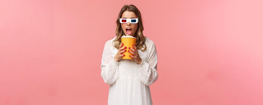 Leisure, going-out and spring concept. Portrait of cute blond funny girl in cinema, eating popcorn from box without hands, looking aside suspicious, wearing 3d glasses, pink background.