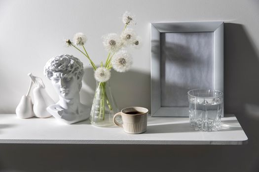 A bunch of fluffy dandelions in a chemical flask, an empty photo frame, a cup of espresso coffee, and a plaster head of David on a white chest of drawers. Copy space