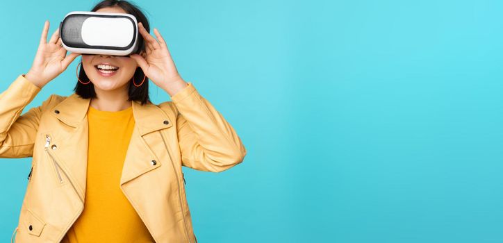 Portrait of happy asian female model using VR headset, smiling and laughing in virtual reality glasses, standing over blue background.
