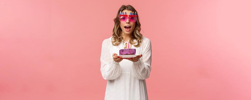 Holidays, spring and party concept. Excited, happy attractive blond woman in white dress, wearing funny b-day glasses, holding birthday cake with lit candle, making wish smiling camera.