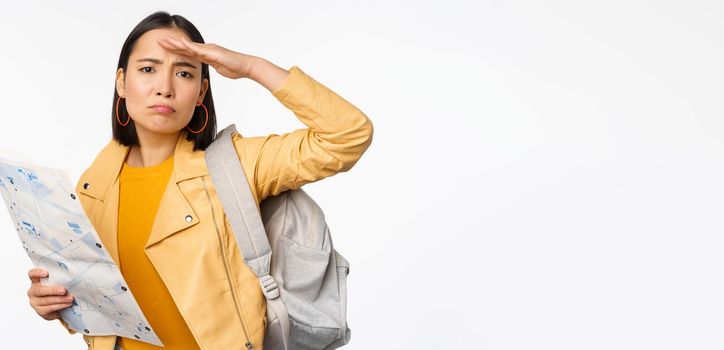 Image of young asian girl tourist, traveller with map and backpack posing against white studio background. Copy space