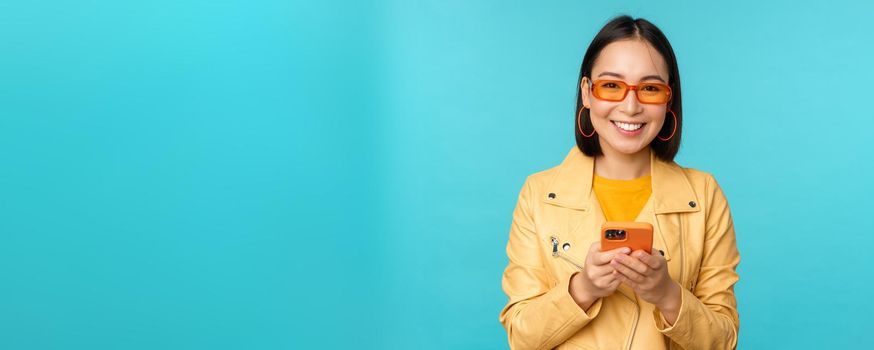 Smiling asian girl in sunglasses, using smartphone app, holding mobile phone, standing over blue background. Copy space