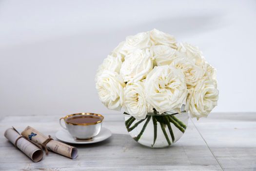 A bouquet of white roses in a round glass vase on a table with a cup of tea and a book. Copy space.