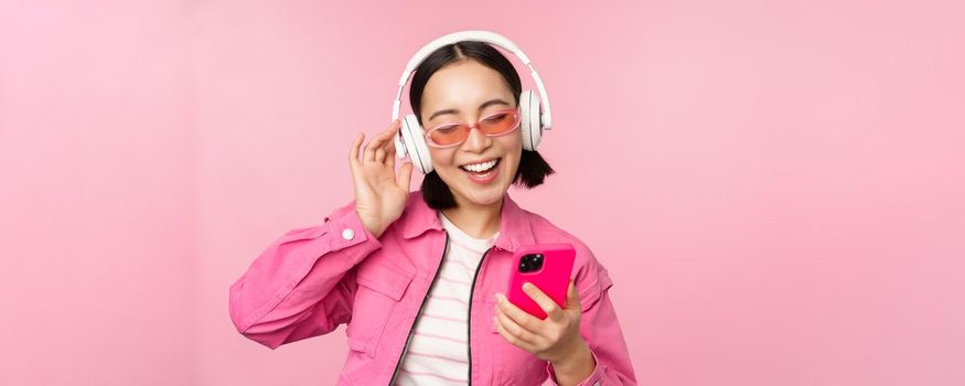 Stylish asian girl dancing with smartphone, listening music in headphones on mobile phone app, smiling and laughing, posing against pink background.