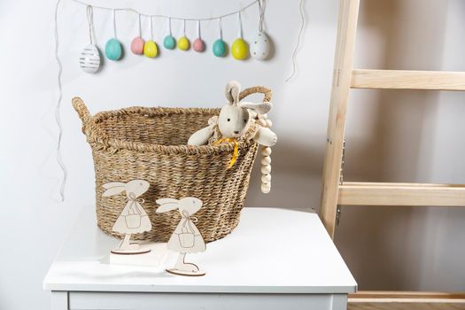 Interior decoration for Easter. A rag rabbit peeks out of a wicker willow basket on the table. A garland of wooden painted eggs above it.