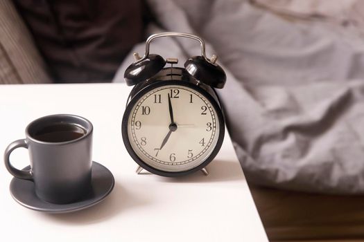 Image alarm clock with a black cup of coffee on a white bedside table in front of the bed with gray linens. The room is in beige tones.