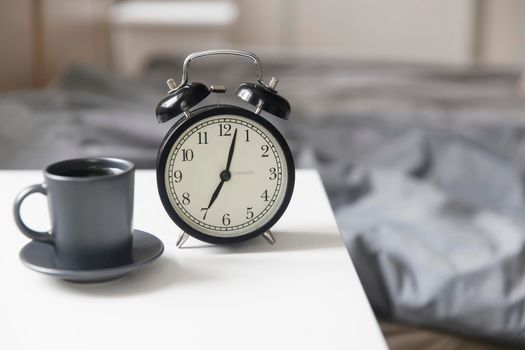 Image alarm clock with a black cup of coffee on a white bedside table in front of the bed with gray linens. The room is in beige tones.