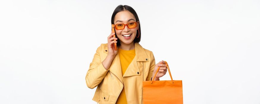 Modern korean girl on shopping, holding store bag, talking on mobile phone and smiling, standing over white background.