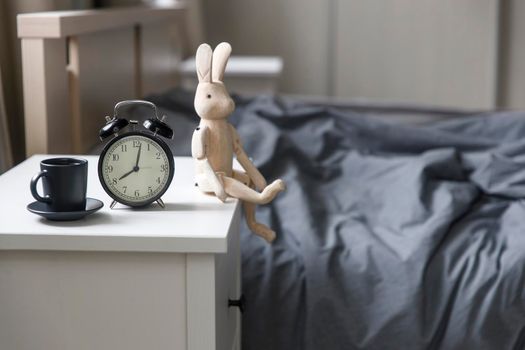 Image alarm clock with a black cup of coffee with figurine of wooden hare on a white bedside table in front of the bed with gray linens. The room is in beige tones.