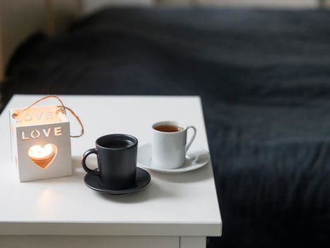 A two cups of coffee: white and black on a tray and a candlestick with the inscription love is on the bedside table