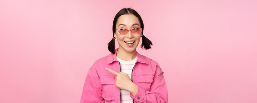 Portrait of smiling asian girl in stylish outfit, sunglasses, pointing finger left, showing advertisement, banner, standing over pink background. Copy space