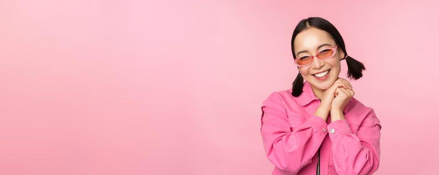 Close up portrait of stylish asian woman in sunglasses, smiling, looking flattered, laughing coquettish, standing over pink background. Copy space