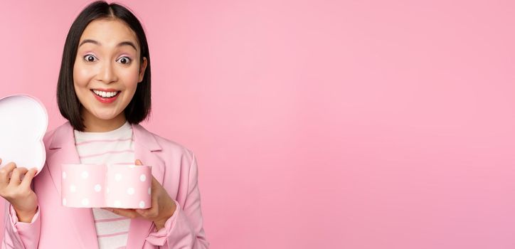 Happy cute korean girl in suit, opens up heart shaped box with romantic gift on white day holiday, standing in suit over pink background.