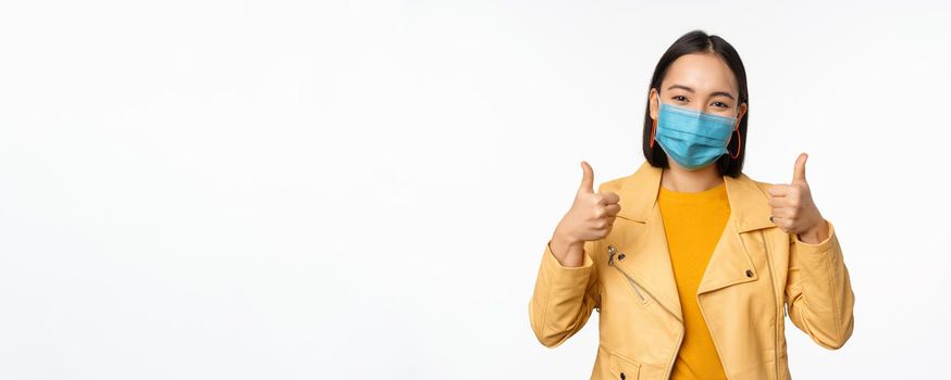 Smiling asian woman in medical face mask showing thumbs up, support wearing protective equipment from covid-19, coronavirus pandemic, white background.