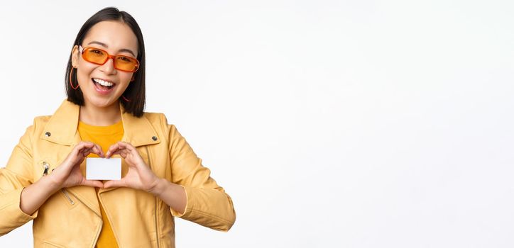 Stylish attractive asian girl in sunglasses, showing credit card and smiling, standing happy against white studio background. Copy space