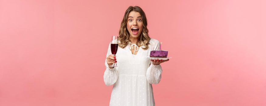 Holidays, spring and party concept. Portrait of fascinated and amused blond girl in white dress look astonished camera, celebrating birthday with b-day cake and glass of wine, pink background.