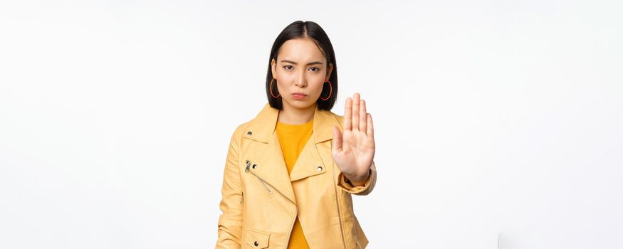 Serious and concerned asian girl extending palm hand, stop gesture, prohibit smth, forbid, standing over white studio background. Copy space