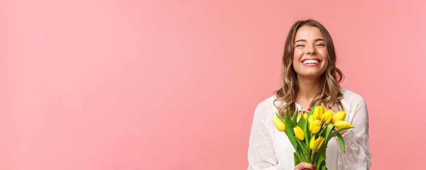 Spring, happiness and celebration concept. Close-up of happy and carefree blond european girl receive beautiful bouquet of flowers, holding yellow tulips, smiling and laughing, pink background.