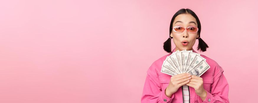 Microcredit and fast loans concept. Excited stylish korean girl, showing money, cash dollars and looking happy, standing in sunglasses over pink background.