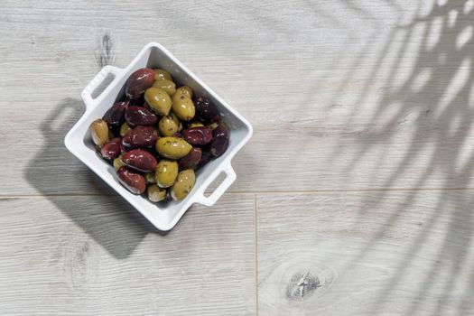 Deep square porcelain bowl with green and dark olives on a wooden board with shadow from a palm tree on a blue background