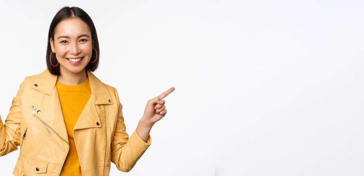 Happy beautiful asian girl pointing fingers left and right, showing banner logo, demonstrating sale, standing in yellow jacket over white background.
