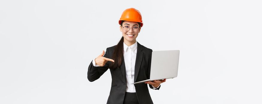 Smiling pleased asian female chief engineer showing chart with enterprise profit, making presentation during investors meeting, wearing safety helmet, pointing at laptop screen, white background.