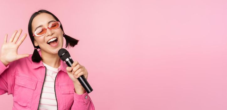 Happy beautiful asian girl singing with mic, using microphone, enjoying karaoke, posing against pink studio background.