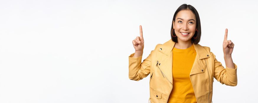 Image of smiling asian brunette woman pointing fingers up, showing advertisement with happy face, posing against white background.