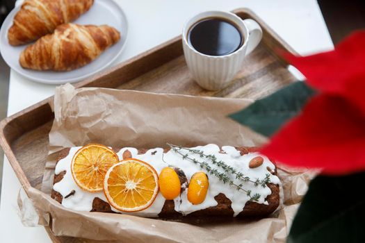 Freshly baked christmas cake and croissant on a gray round plate, a white cup of coffee and a garland on a tray on the table. Blooming poinsettia.