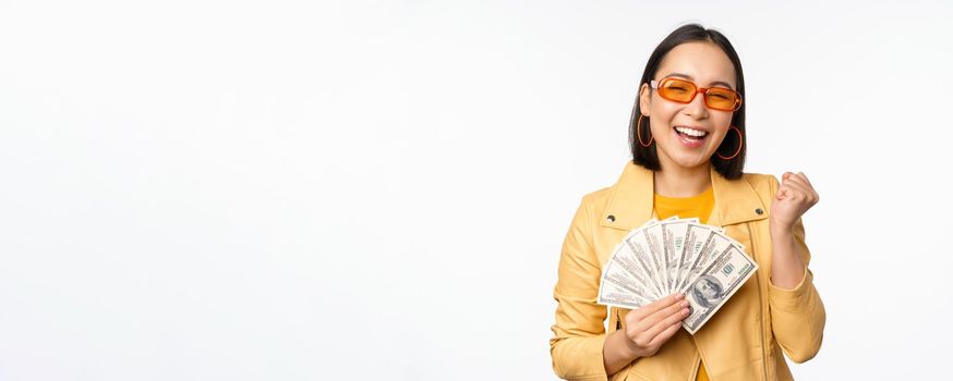 Stylish smiling asian girl holding money cash, showing dollars and celebrating, standing over white background. Copy space