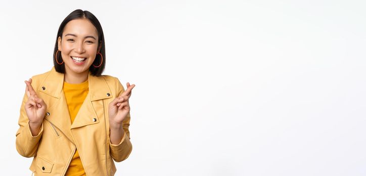Portrait of excited asian woman looks hopeful, wishing, praying or begging, waiting for news, standing over white background, smiling enthusiastic.
