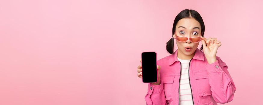 Portrait of asian girl showing mobile phone screen, reacting surprised, standing over pink background. Copy space