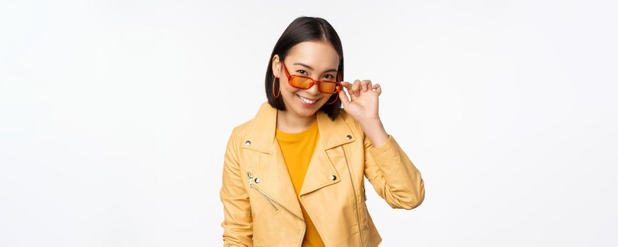 Beautiful asian girl in sunglasses, smiling and looking happy, posing in yellow jacket against white background. Copy space