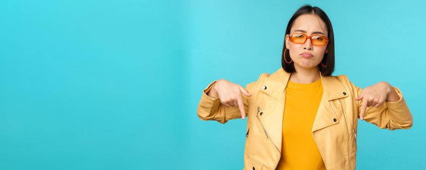 Sad and gloomy chinese girl in sunglasses, points fingers down and grimaces, looks miserable and disappointed, emotion of regret, standing over blue background.