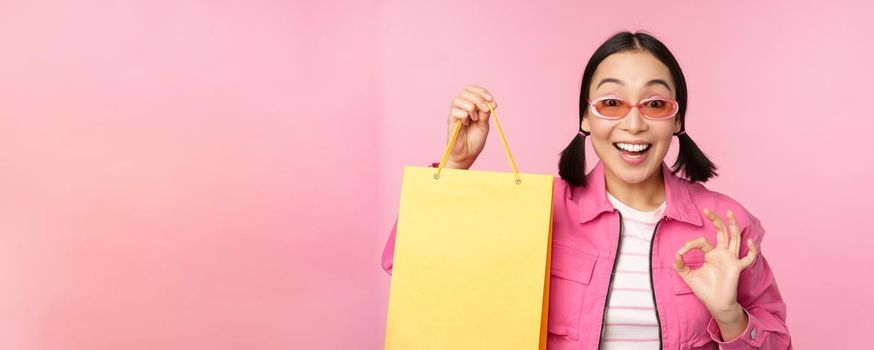Shopping. Stylish asian girl in sunglasses, showing bag from shop and smiling, recommending sale promo in store, standing over pink background.