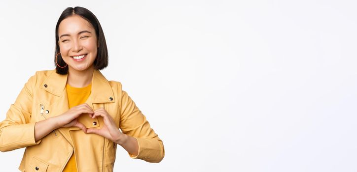 Image of beautiful asian girl showing heart, love sign, express care and romantic feelings, standing over white studio background.