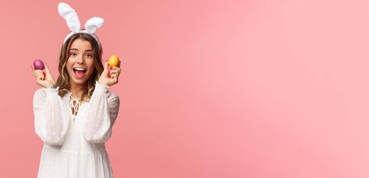 Holidays, spring and party concept. Portrait of excited charming young woman celebrating Easter in rabbit ears and white party dress, dancing with two painted eggs, pink background.