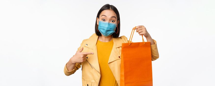 Covid-19 and shop concept. Young asian stylish woman, wearing medical face mask, holding shopping bag, going in malls during pandemic, white background.