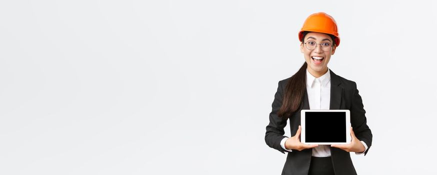 Excited and amazed smiling asian female construction engineer, architect in safety helmet and business suit, showing digital tablet screen with amazed expression, standing white background.