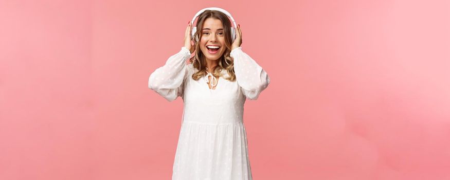 Portrait of excited, happy good-looking girl in white tender dress, wearing headphones and smiling amazed as looking at camera, fascinated with good sound quality, pink background.