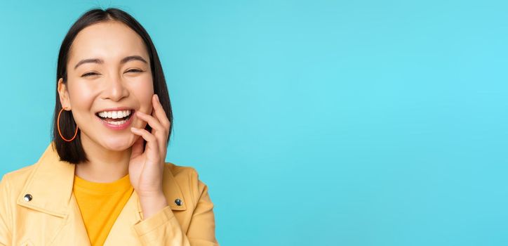 Close up portrait of natural asian girl laughing, smiling and looking happy, standing over blue background. Copy space