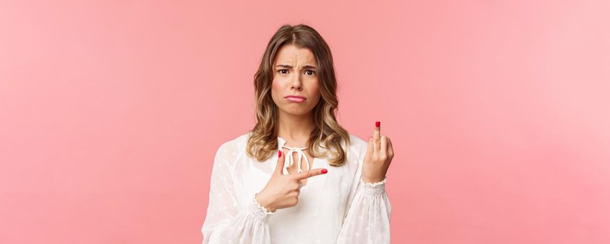 Close-up portrait sad and frustrated young blond girl waited for proposal during romantic date, pointing at finger without weddint ring with puzzled upset expression, standing pink background.