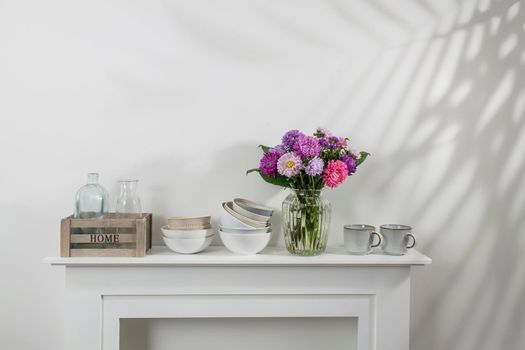 kitchen utensils, cups, bowls, bottles, storage items, a bouquet of asters are on a white dresser. Copy space