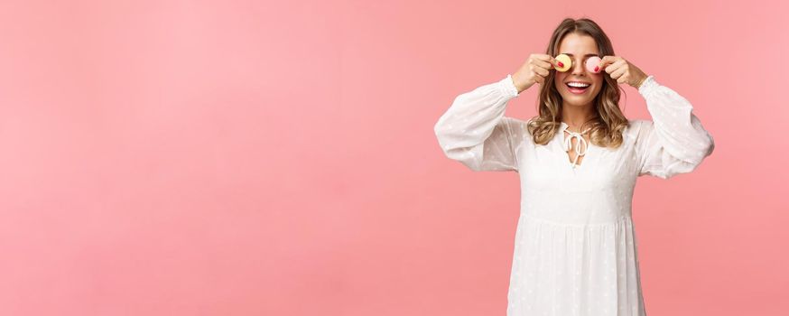 Holidays, spring and party concept. Portrait of carefree funny and cute blond girl in white dress, holding two macarons on eyes and smiling happy, like eating tasty desserts, pink background.