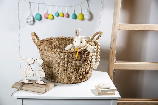 Interior decoration for Easter. A rag rabbit peeks out of a wicker willow basket on the table. A garland of wooden painted eggs above it.