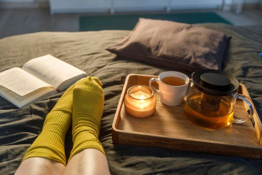 Soft photo of woman`s legs in the woolen socks on the bed with book and cup of tea and candle on the tray. Interior and home cosiness concept. Top view