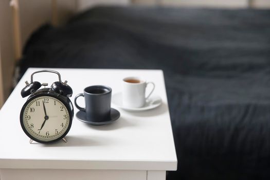 Image alarm clock with a black cup of coffee on a white bedside table in front of the bed with gray linens. The room is in beige tones.