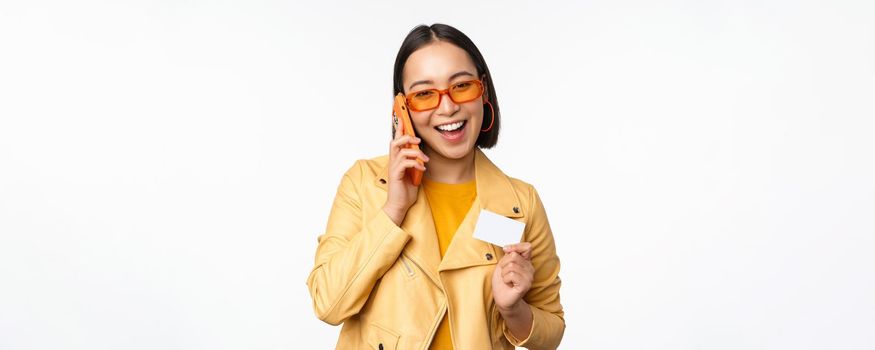 Stylish asian female model, talking on smartphone and showing credit card, standing over white background. Copy space