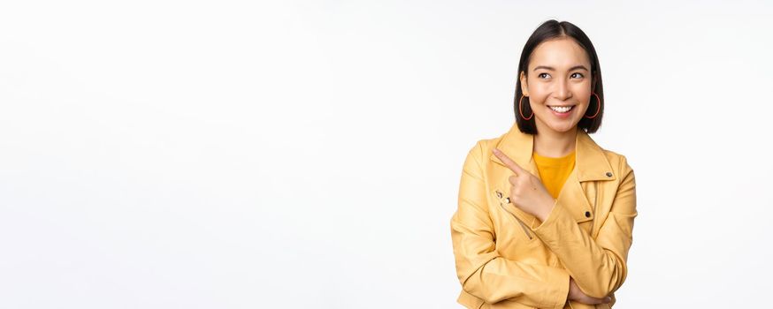 Portrait of smiling asian woman pointing and looking left, choosing item, showing logo or banner, standing over white background.