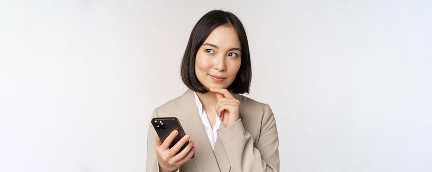 Close up portrait of korean woman, corporate lady in suit, using mobile phone and smiling, holding smartphone, standing over white background.